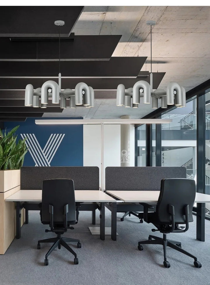 Suspension design tubulaire aux courbes en U en finition gris mat installée au-dessus d'un bureau moderne, offrant un éclairage fonctionnel et un style contemporain.