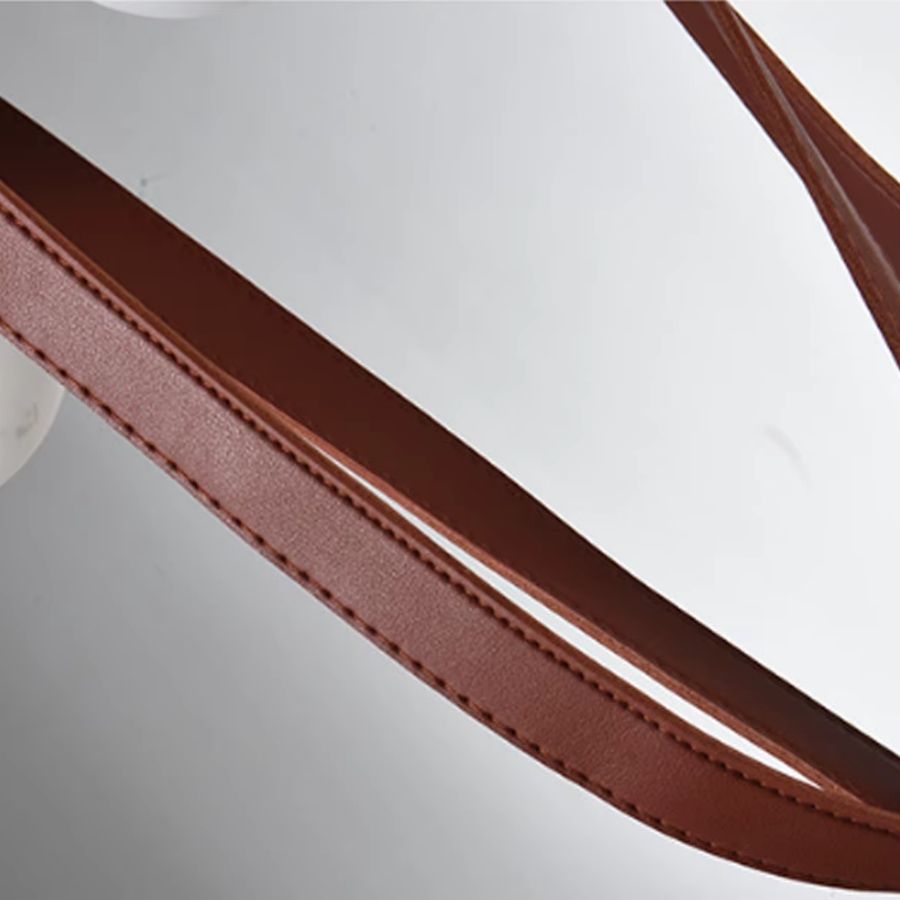 Détail d’une lanière en cuir brun aux coutures apparentes, soulignant le caractère artisanal et sculptural du luminaire.