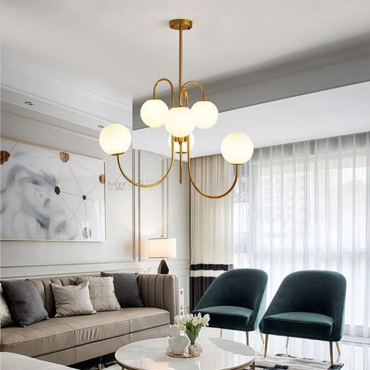 Éclairage suspendu moderne en laiton et boules de verre opalin dans un salon lumineux avec fauteuils verts et canapé beige.