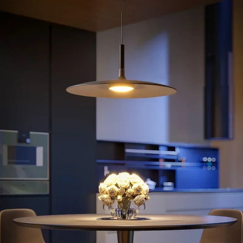 Luminaire design en forme de disque et aile évasée éclairant une table ronde en bois clair dans une cuisine chaleureuse.