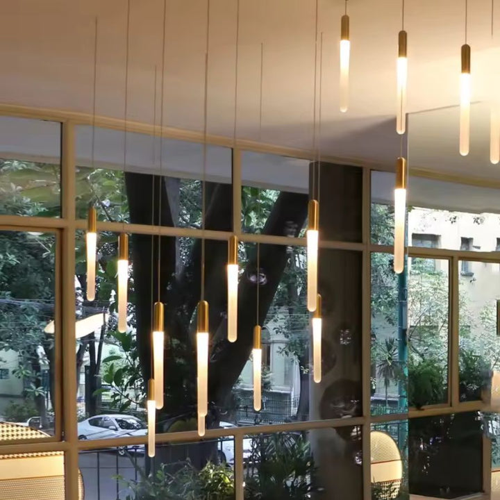 Sous une grande verrière, ce luminaire tubulaire doré déploie ses diffuseurs allongés dans une cascade contemporaine