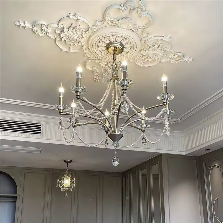 Lustre argenté d'inspiration baroque orné de pampilles de cristal et de perles, sous un plafond à moulures sculptées.