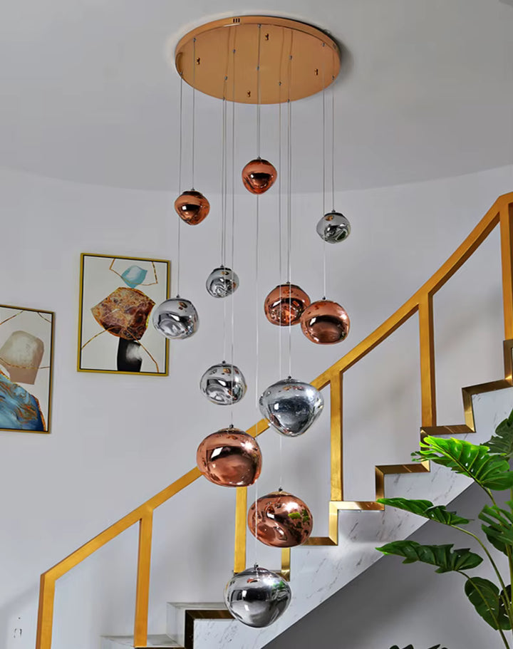 Une suspension design aux sphères cuivrées et chromées effet miroir descend en cascade près d'un escalier à rampe dorée.
