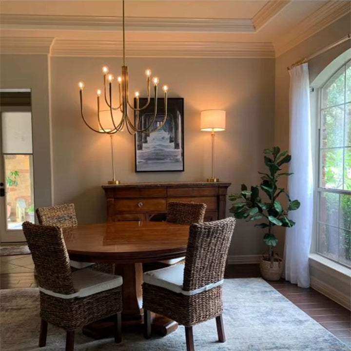 Au-dessus d'une table ronde en bois, lustre doré à bras effilés et lumières façon bougies, au style moderne et épuré.