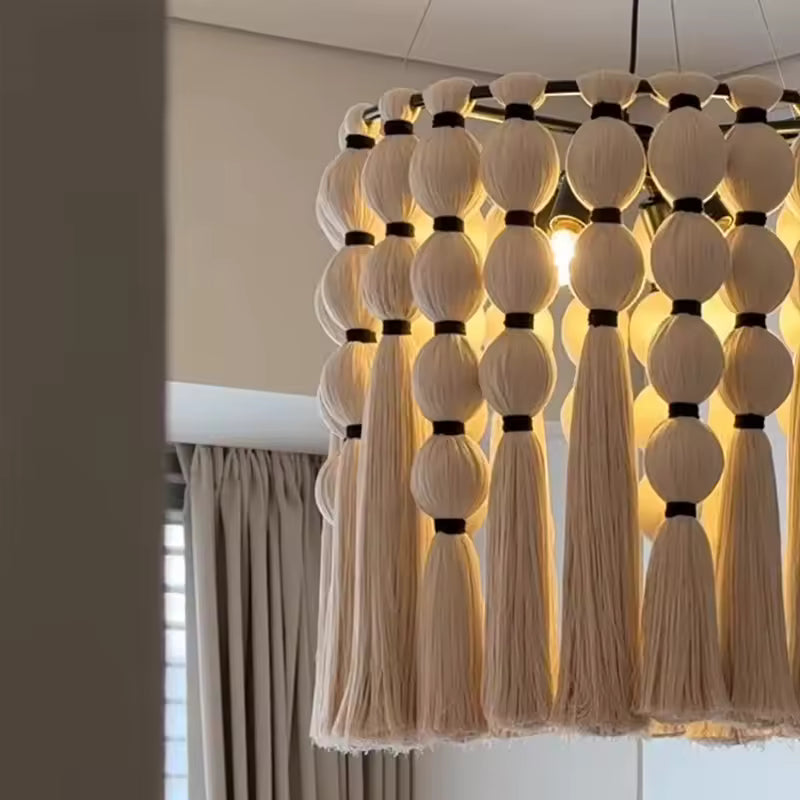 Vue rapprochée d'un lustre macramé en coton écru, aux pompons et franges bohèmes illuminés d'une lumière dorée.