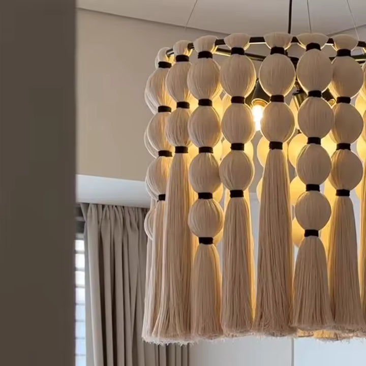 Vue rapprochée d'un lustre macramé en coton écru, aux pompons et franges bohèmes illuminés d'une lumière dorée.