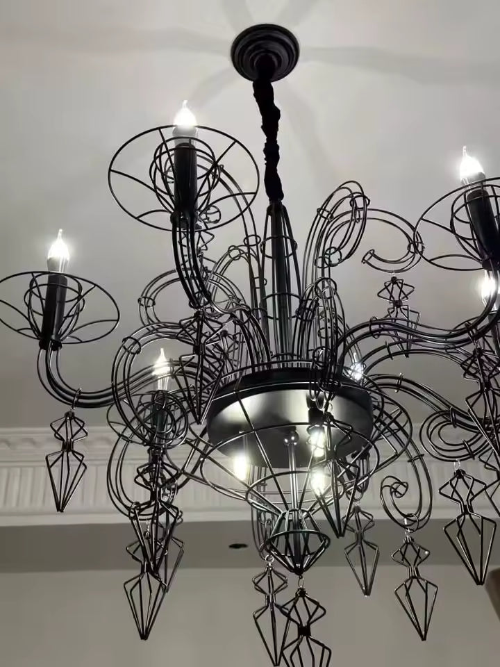 Vue rapprochée d'un lustre noir baroque illuminé, aux volutes filaires et pampilles géométriques, d'inspiration néogothique.