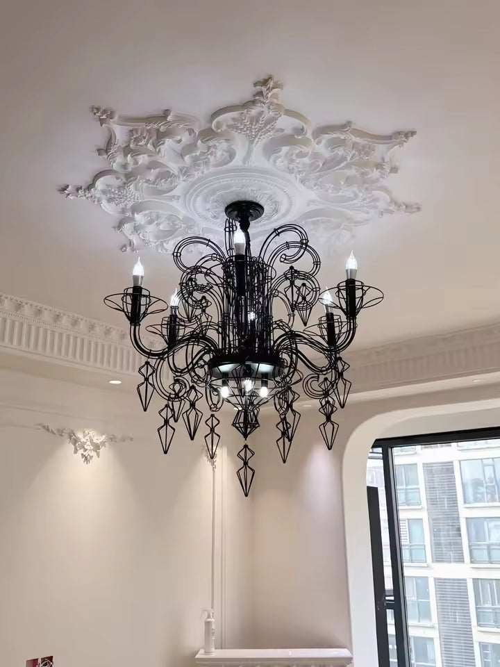 Dans un intérieur haussmannien, lustre noir baroque à volutes filaires et pampilles géométriques sous une rosace ouvragée.
