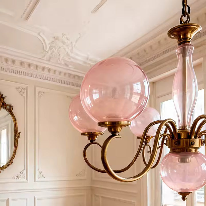 Détail d’un lustre rose à globes en verre et bras dorés courbés, dans un intérieur classique aux accents baroques