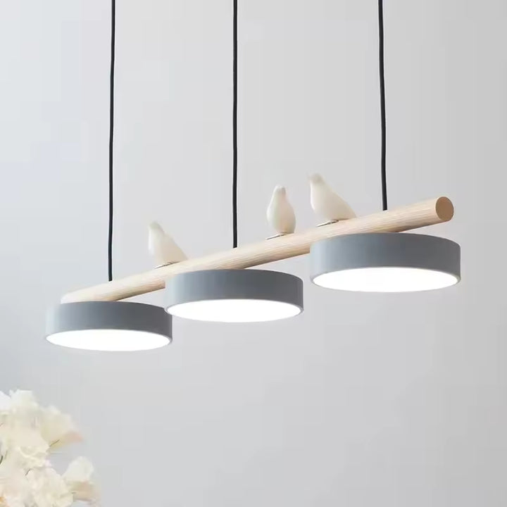 Trois disques lumineux suspendus sous une barre en bois ornée d’oiseaux, dans un style scandinave poétique