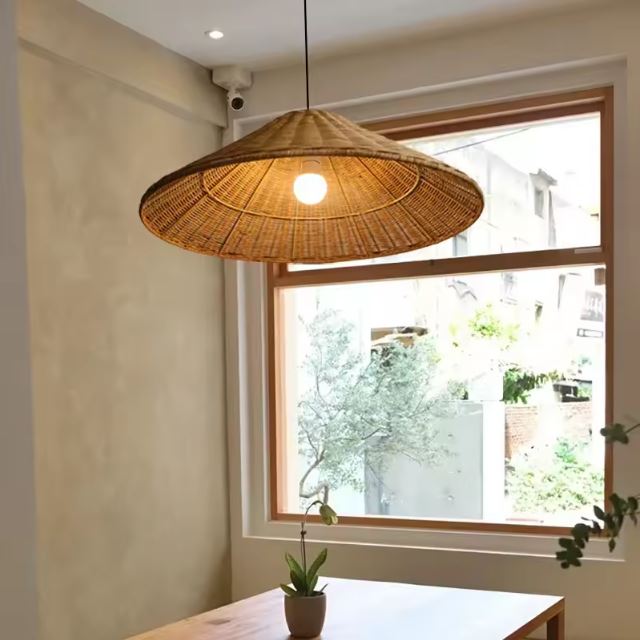 Suspension chapeau en rotin naturel éclairant une table en bois près d'une fenêtre avec vue sur un jardin.