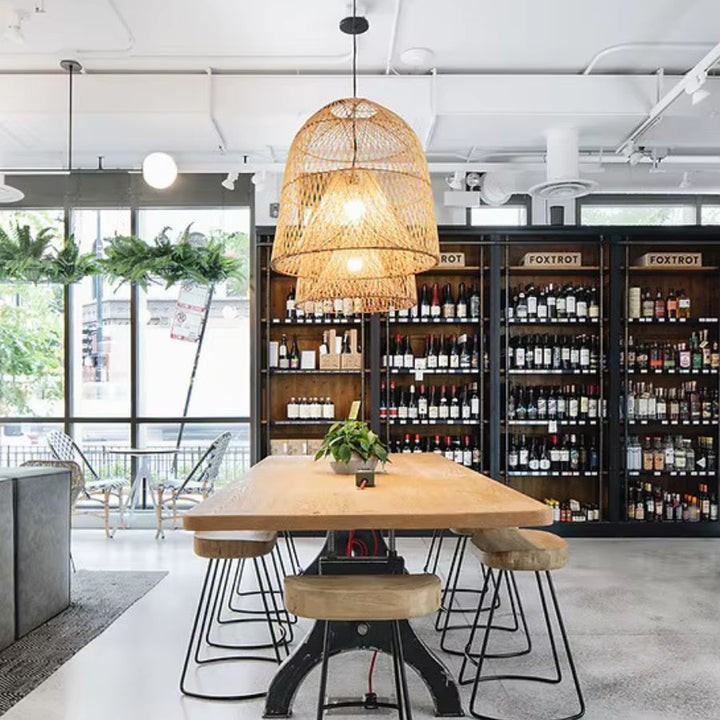 Grande suspension en rotin tressé à double cloche ajourée, surplombant une table haute dans un espace convivial au style industriel.