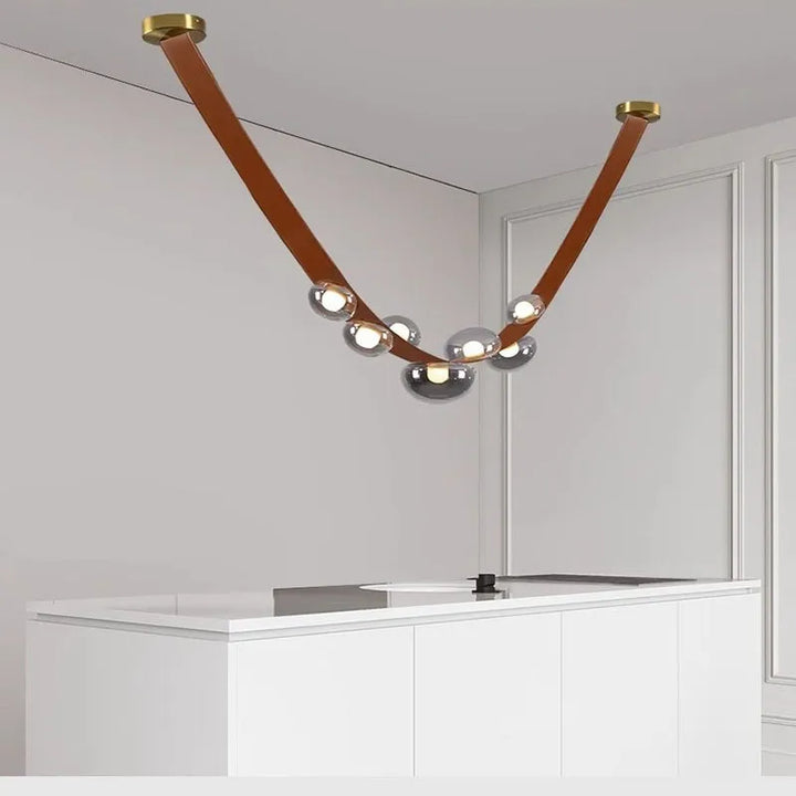 Luminaire suspendu chic en cuir marron cognac et sphères de verre fumé, installé au plafond d'une salle à manger lumineuse.