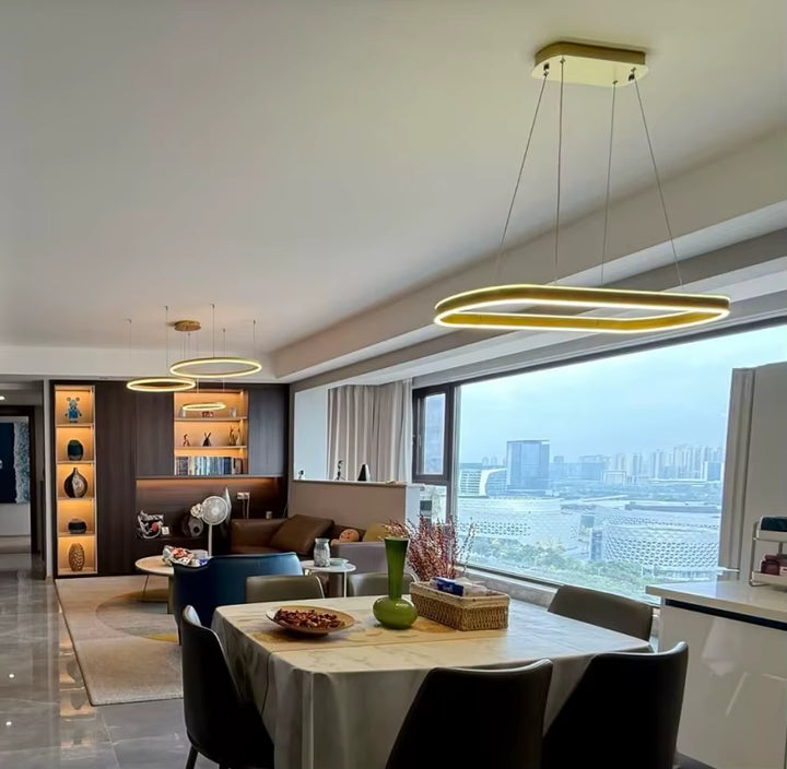 Près d'une baie vitrée, suspension LED dorée à anneau ovale allongé, au style contemporain chic, surplombant une table à manger.
