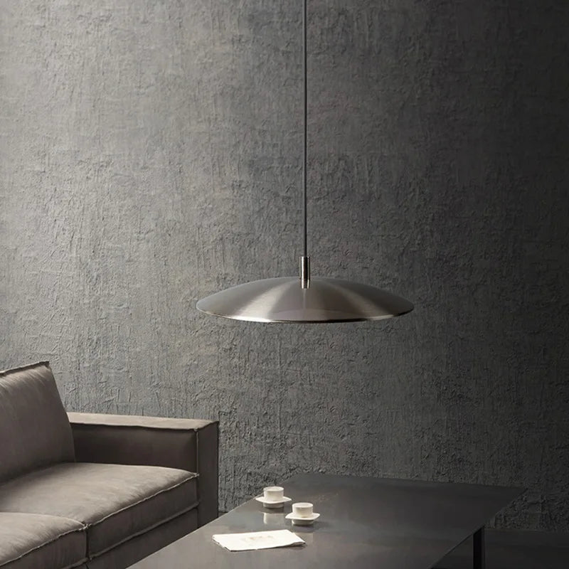 Suspension disque design en métal argenté installée dans un salon épuré avec un mur effet béton.
