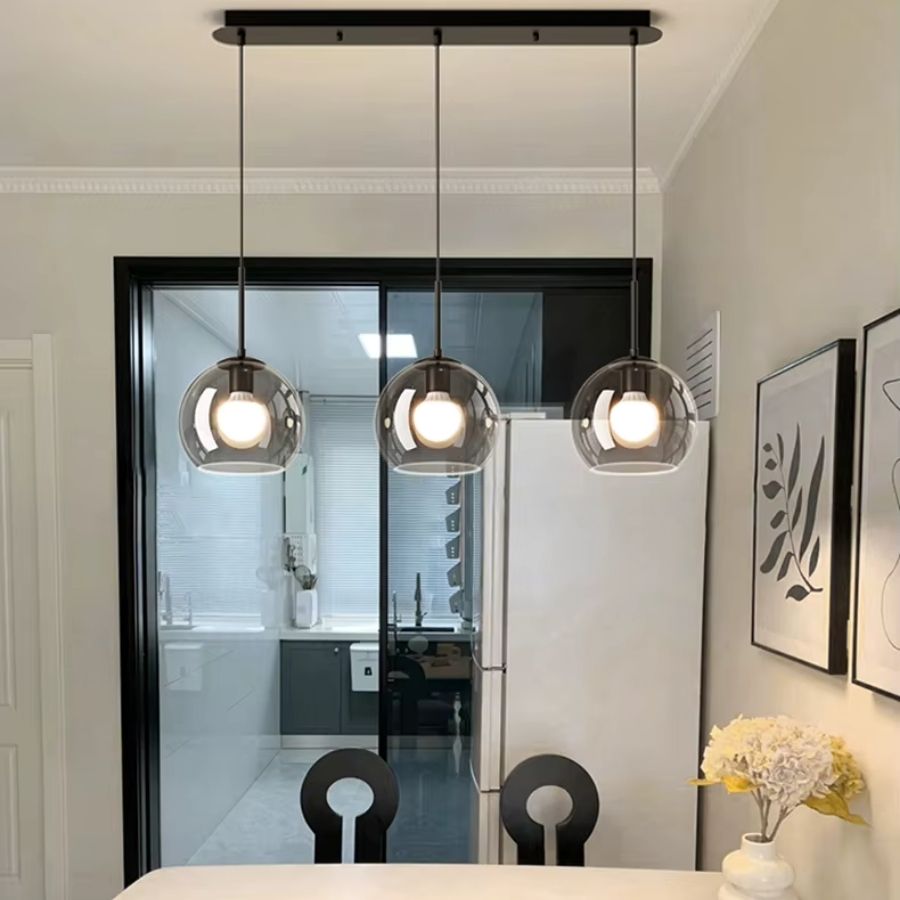 Installé au-dessus de la table, ce luminaire en métal noir à trois globes fumés en verre renforce l’élégance contemporaine de la pièce