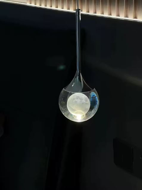 Suspension goutte en verre transparent et métal noir sur fond sombre, luminaire design contemporain à l’élégance raffinée.