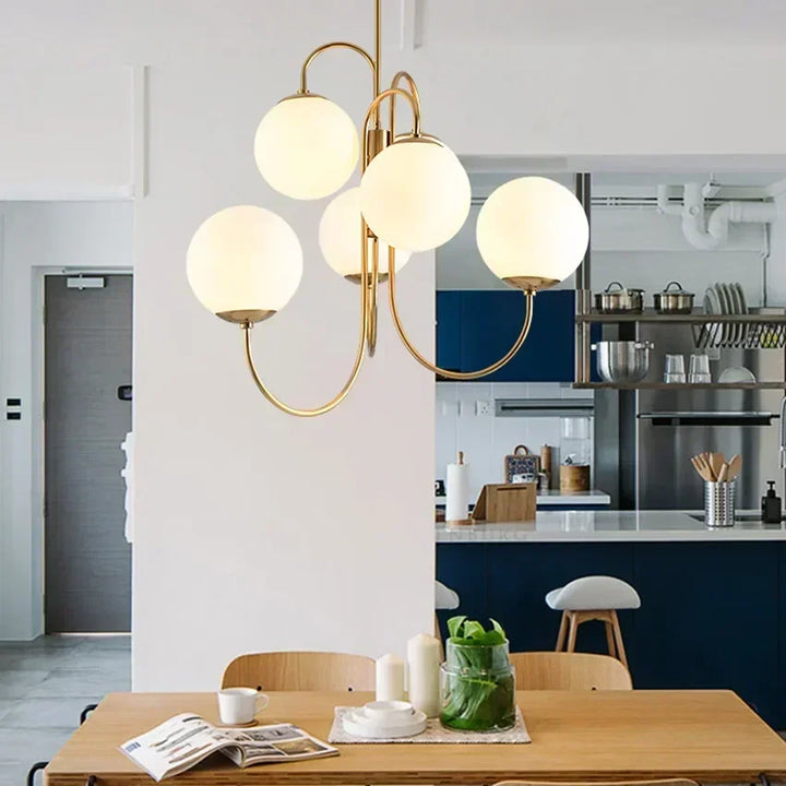 Suspension design en laiton doré avec boules en verre opalin blanc, éclairant une cuisine ouverte avec coin repas.
