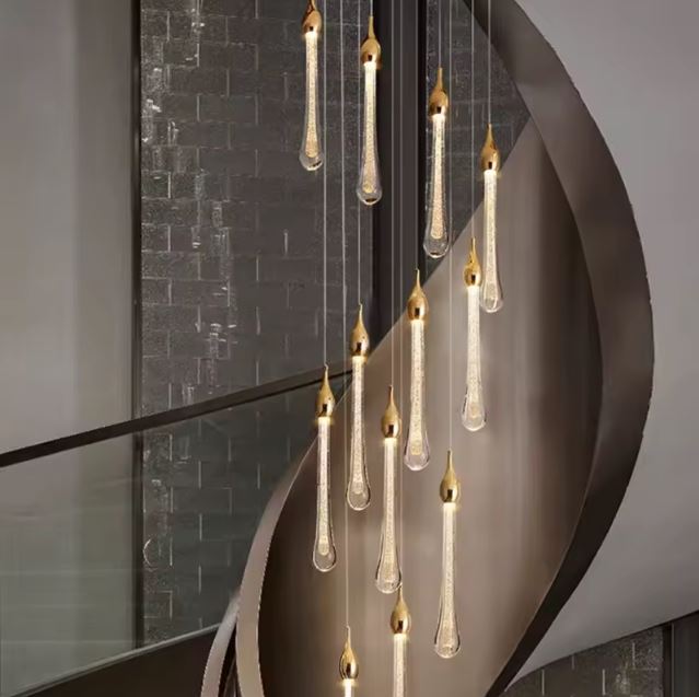 Suspension LED dorée en forme de gouttes installée près d’un escalier colimaçon avec mur effet brique noire et ambiance design.