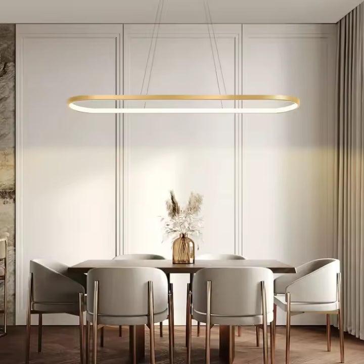 Au-dessus d’une table de salle à manger, la suspension LED dorée dessine un cadre lumineux aux lignes minimalistes