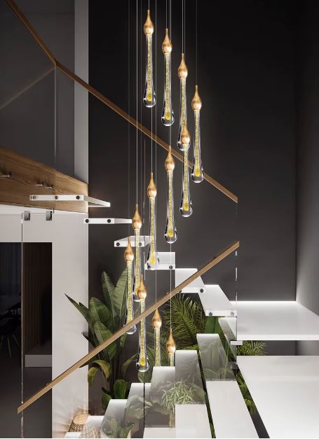 Suspension goutte LED dorée éclairant un escalier flottant design avec garde-corps en verre et végétation tropicale en fond.