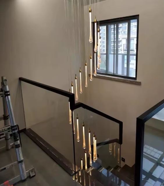 Suspension goutte LED dorée installée dans une cage d’escalier à double hauteur avec fenêtre vitrée et garde-corps en verre noir.