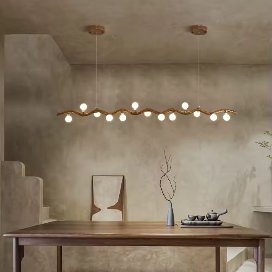 Dans une salle à manger wabi-sabi aux murs texturés, une longue suspension effet bois déploie ses globes lumineux.