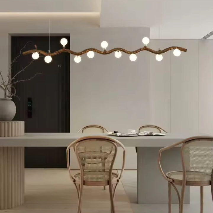 Au-dessus d'une table aux chaises en cannage, suspension à branche ondulée effet bois et globes lumineux, au style japandi.