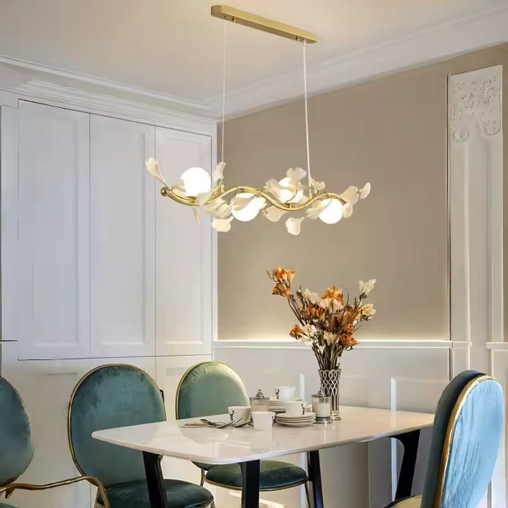 Dans une salle à manger élégante aux fauteuils verts, suspension florale dorée à globes opalins et fleurs blanches.
