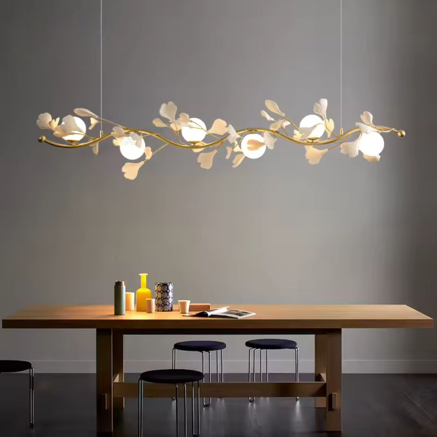 Une suspension florale linéaire aux rameaux dorés et globes opalins surplombe une table en bois dans un décor minimaliste.