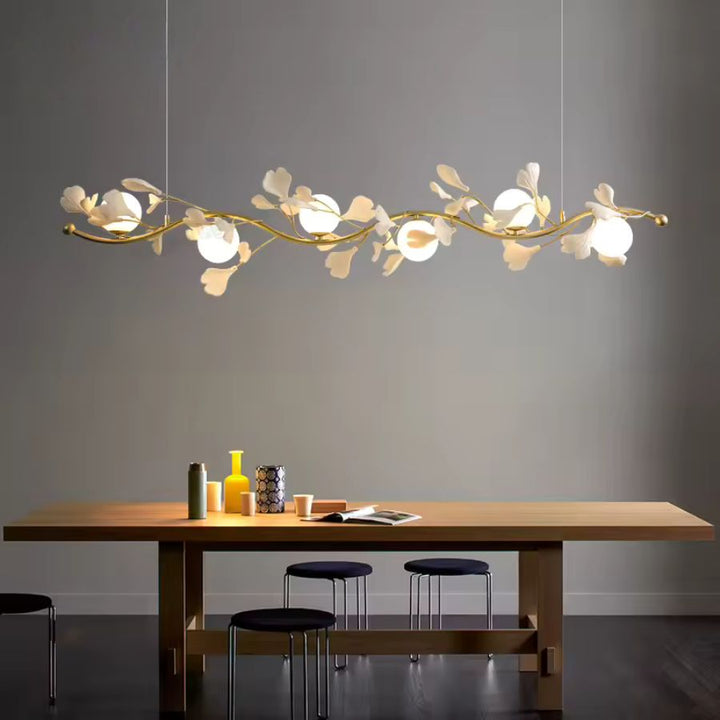 Une suspension florale linéaire aux rameaux dorés et globes opalins surplombe une table en bois dans un décor minimaliste.