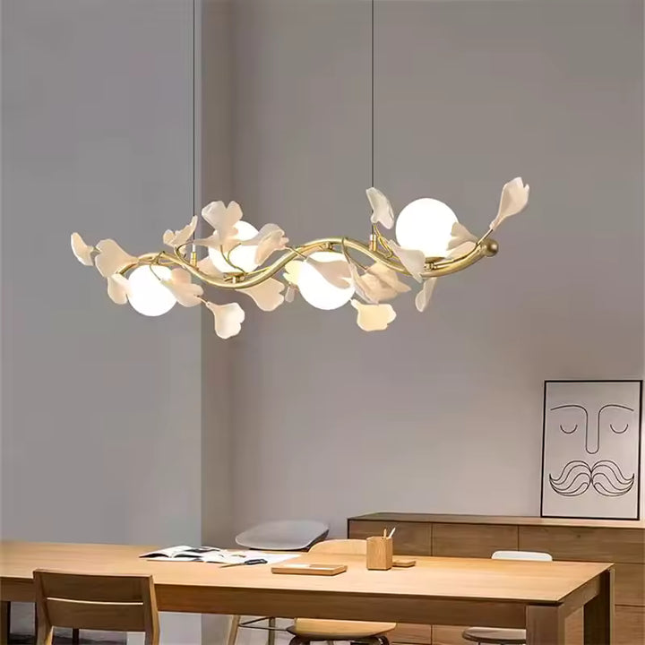 Au-dessus d'une table en bois, suspension linéaire florale aux branches dorées, fleurs blanches et globes opalins, de style organique.