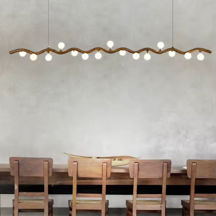 Une suspension en forme de branche ondulée à globes lumineux surplombe une longue table en bois au style naturel épuré.