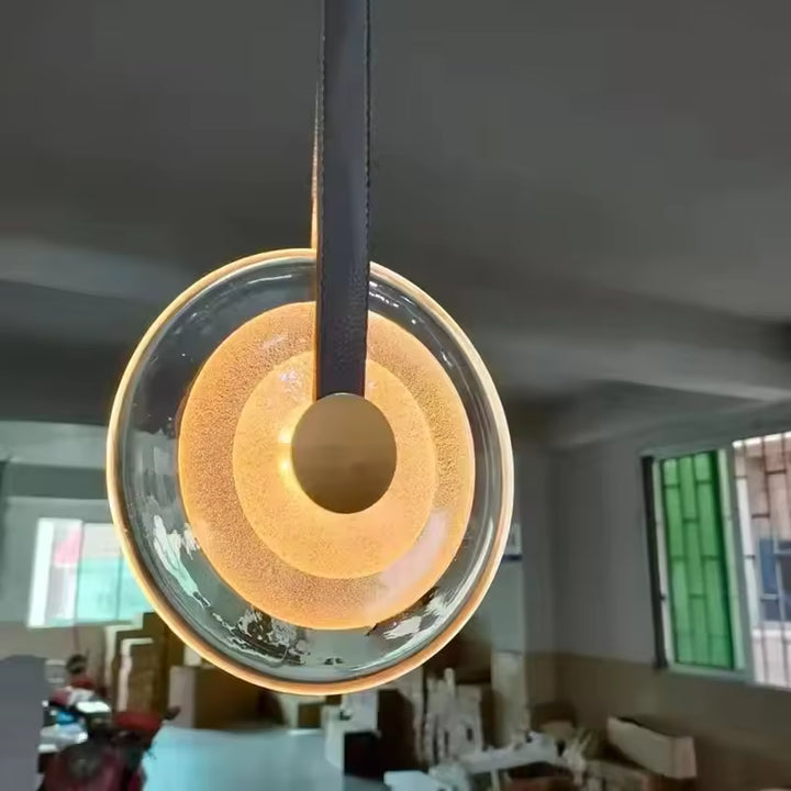 Suspension médaillon en verre avec lanière en cuir, diffusant un halo chaleureux dans un style moderne et raffiné