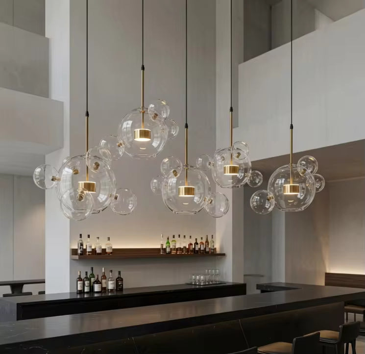 Cinq suspensions nuage de bulles en verre soufflé transparent et détails dorés surplombent un bar moderne à haut plafond.