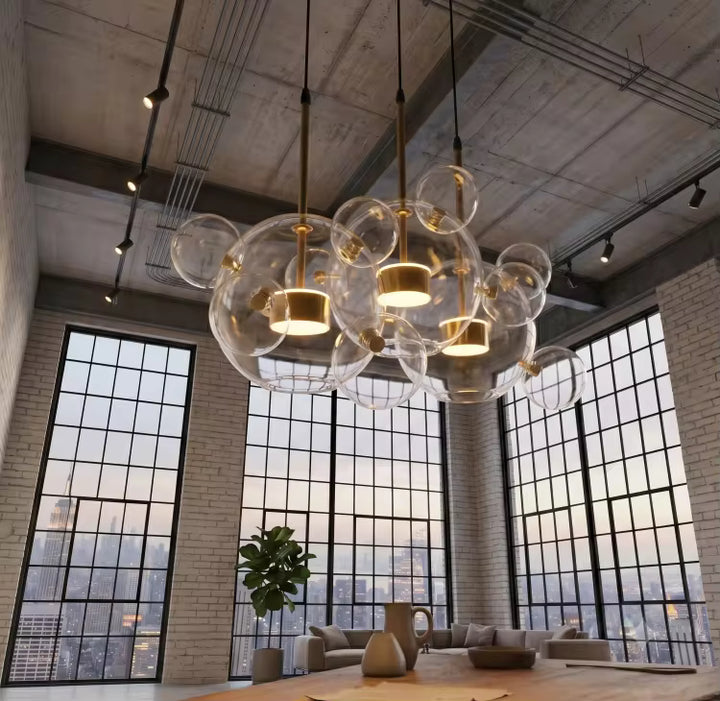 Trois suspensions à bulles en verre soufflé transparent illuminent un loft industriel aux grandes verrières et au plafond brut.