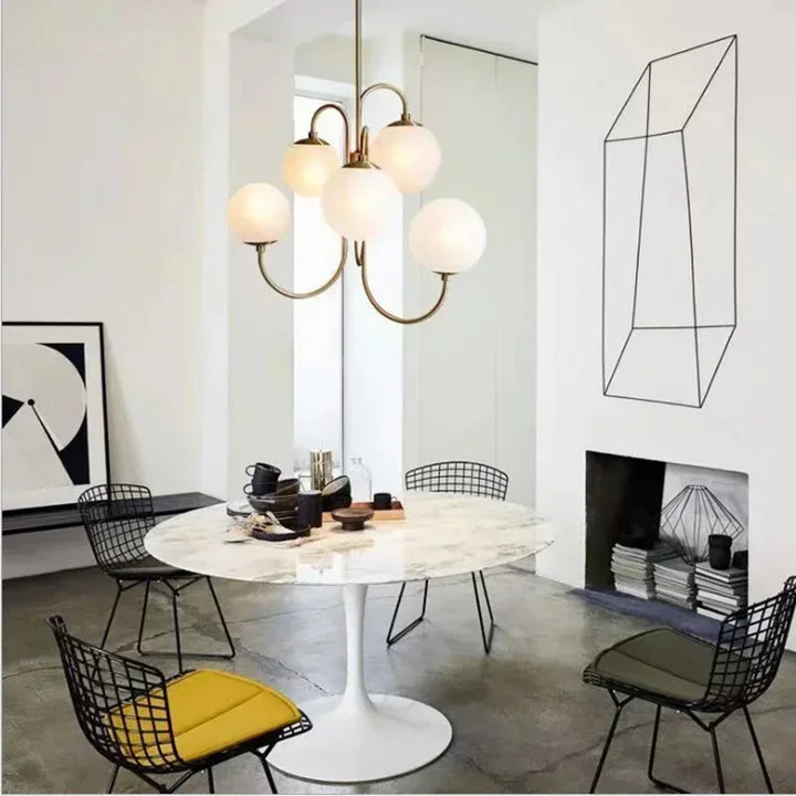 Suspension design en laiton et opaline au-dessus d'une table de salle à manger ronde avec chaises filaires noires et décor minimaliste.