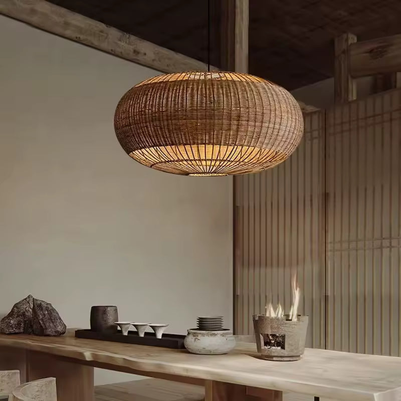 Une suspension ovale en rotin tressé diffuse une lumière douce au-dessus d'une table en bois, dans un décor rustique et japandi.