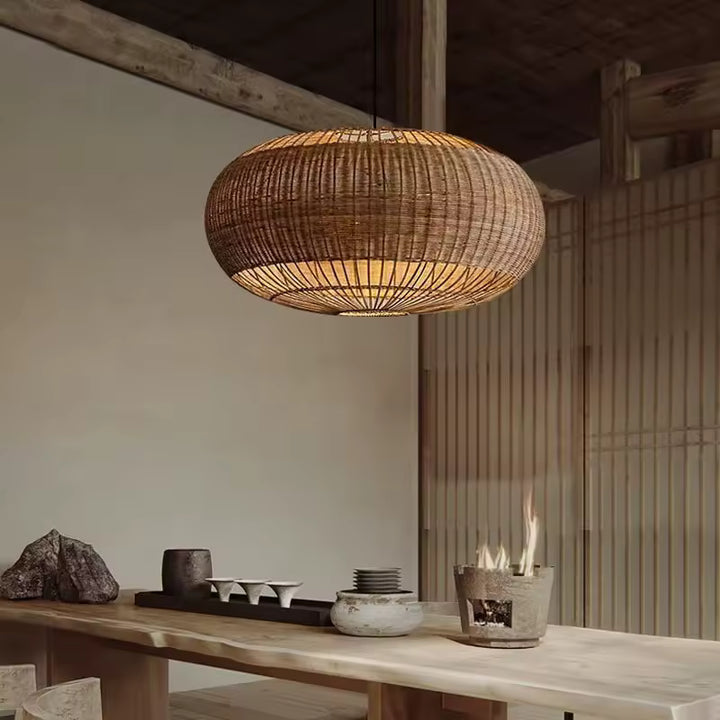 Une suspension ovale en rotin tressé diffuse une lumière douce au-dessus d'une table en bois, dans un décor rustique et japandi.