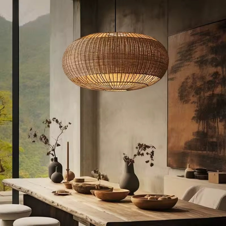 Au-dessus d'une table en bois brut, suspension ovale en rotin tressé ajouré, dans un intérieur au style wabi-sabi.