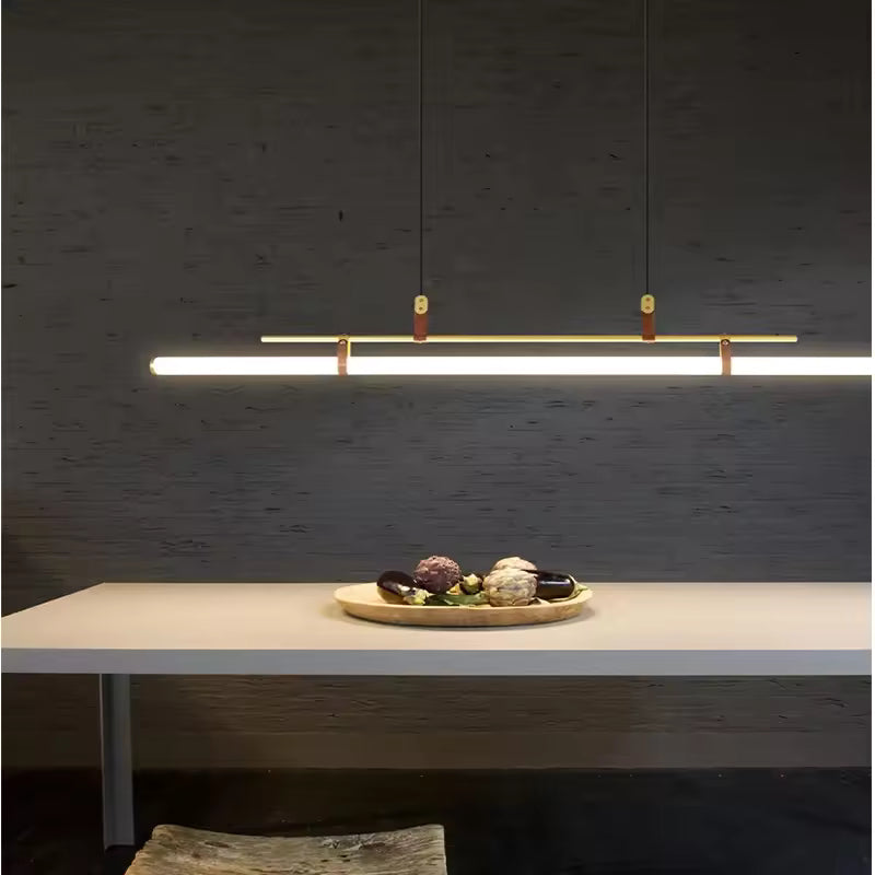 Suspension tube LED dorée avec attaches en cuir, installée au-dessus d’un îlot dans une cuisine minimaliste