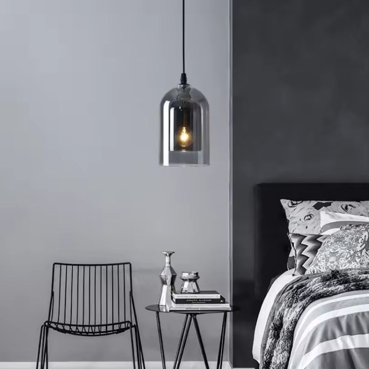 Une suspension en verre fumé à finition chromée éclaire un coin de chambre au style industriel minimaliste, près d'un fauteuil noir.