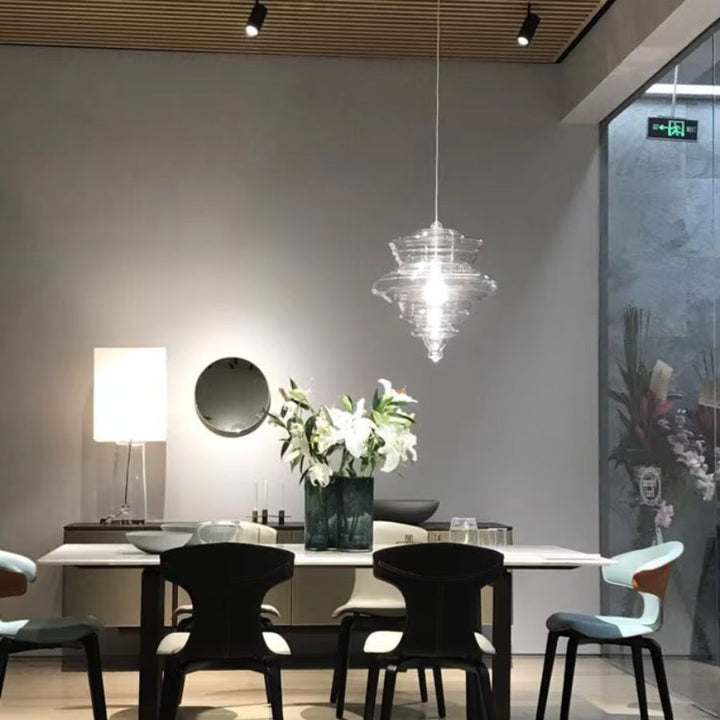 Suspension en verre soufflé transparent aux reliefs sculpturaux étagés, illuminant une table dans une salle à manger moderne.