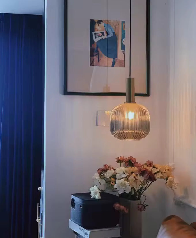 Suspension en verre cannelé fumé avec finition dorée installée dans un coin de salon moderne, à proximité d’un bouquet de fleurs et d’un cadre mural. Ce luminaire diffuse une lumière chaleureuse et crée une ambiance élégante et accueillante.