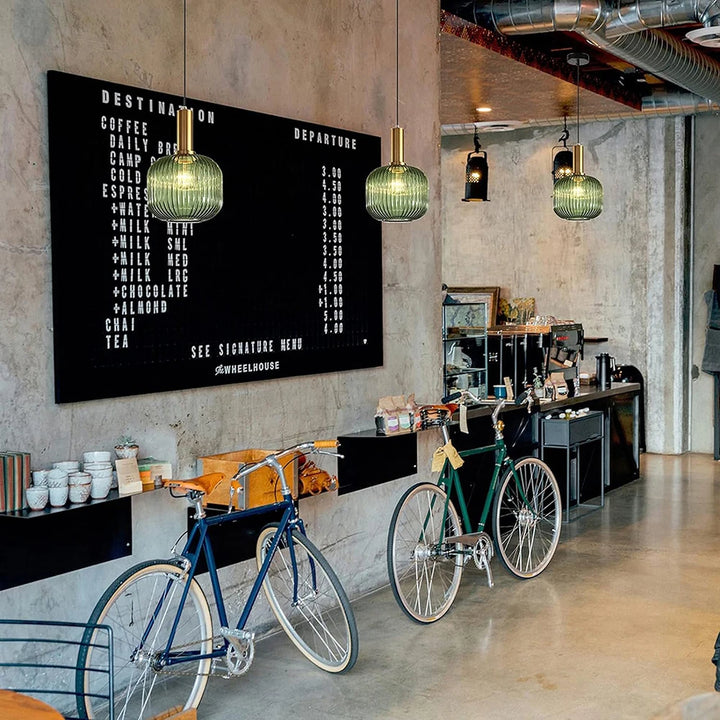 Suspensions en verre cannelé vert avec finitions dorées installées dans un café au style industriel. Ces luminaires décoratifs apportent une ambiance chaleureuse et élégante à l’espace de restauration