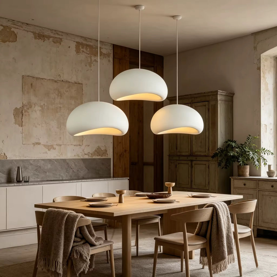 Trois suspensions blanches aux dômes asymétriques diffusent une lumière chaleureuse sur une table en bois, dans un décor campagne chic.