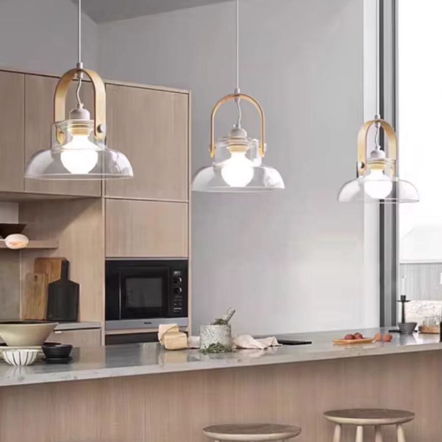 Trois suspensions en bois clair et verre soufflé transparent habillent un îlot de cuisine lumineux au style moderne et naturel