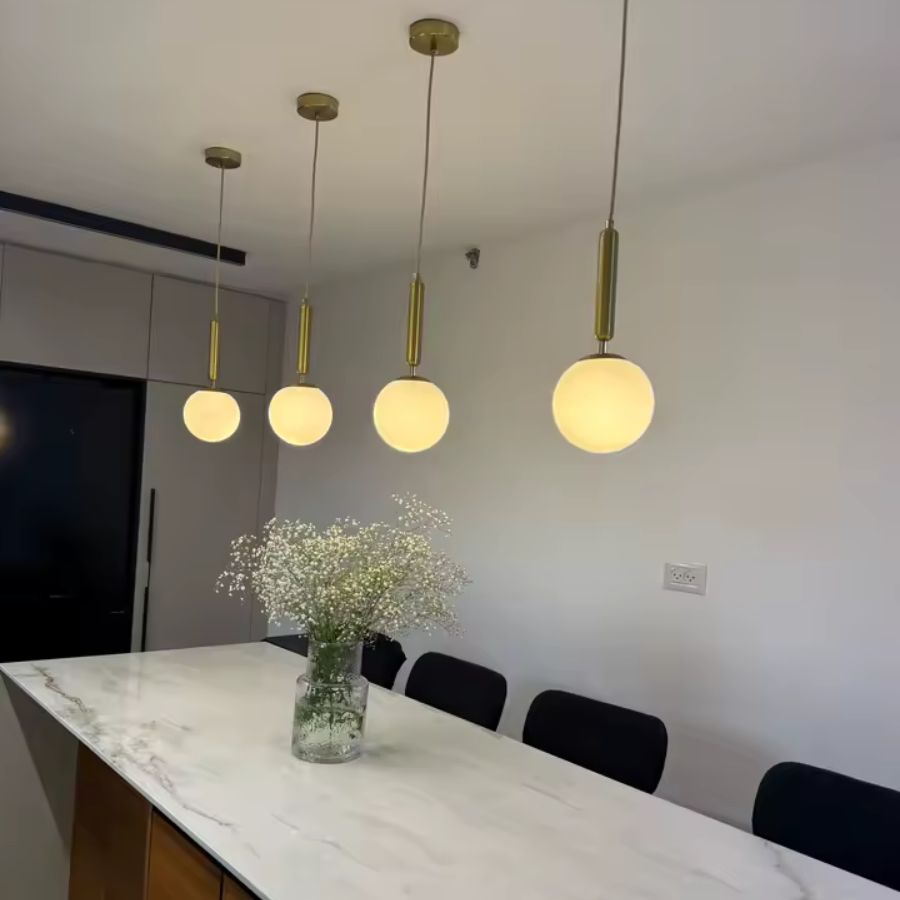 Alignement de suspensions globe blanches à tiges dorées au-dessus d’un îlot, dans une cuisine moderne et épurée
