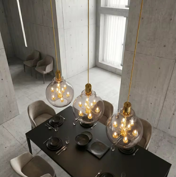 Trois suspensions globe en verre transparent avec lustres dorés, installées au-dessus d’une table sous plafond haut