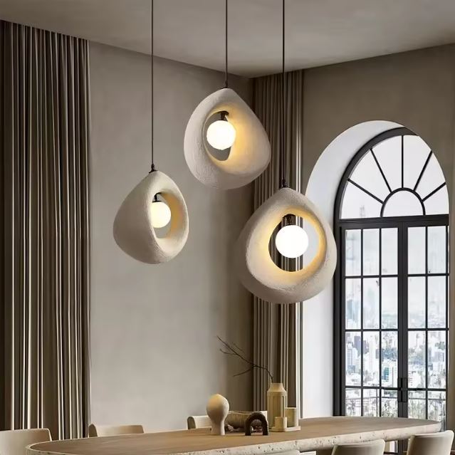 Trois suspensions en plâtre au design organique éclairant une table moderne dans une pièce avec une grande fenêtre en arche.